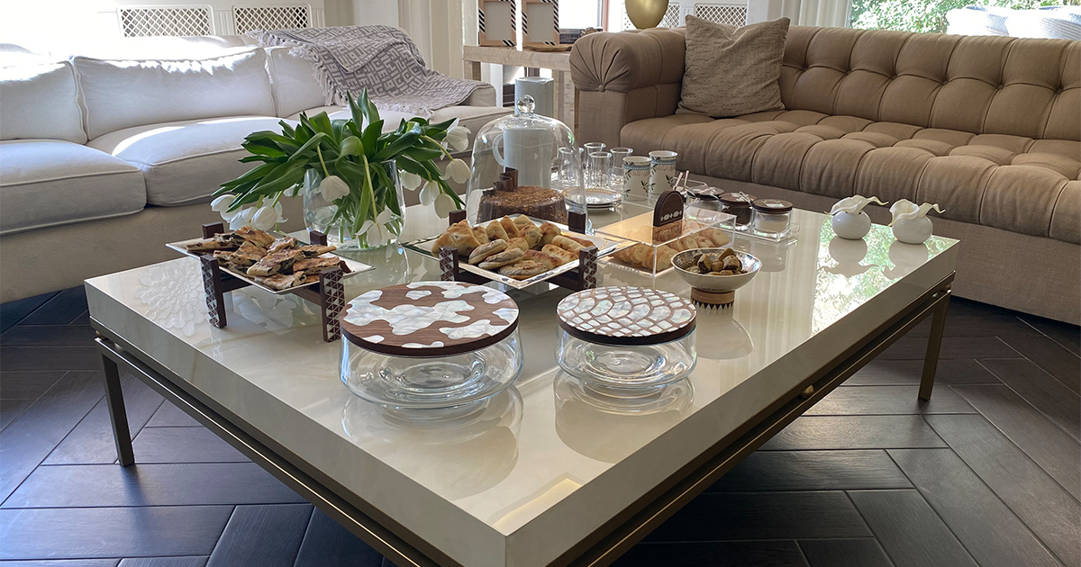The alt text for the image you sent is:

A coffee table in a living room, styled with oriental touches for a morning coffee ritual. The table is adorned with an acrylic tray with mother-of-pearl handles, an olive mug, matching dessert plates, a porcelain thermos, a tiny vase with a white flower, and aluminum plates with mother-of-pearl designs elevated on wooden risers.